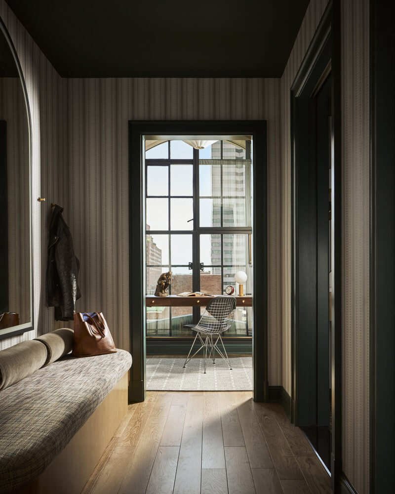 A hallway with striped wallpaper leads to a windowed door opening to a small patio with a table, chair, and city view. A coat hangs by a cushioned bench with a brown bag on it.