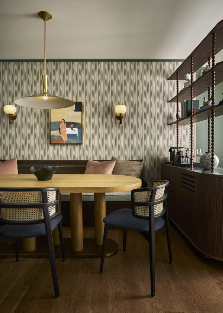 A dining area with a wooden oval table, four chairs, patterned wallpaper, a wall painting, built-in bench seating, and a shelving unit with decorative items.