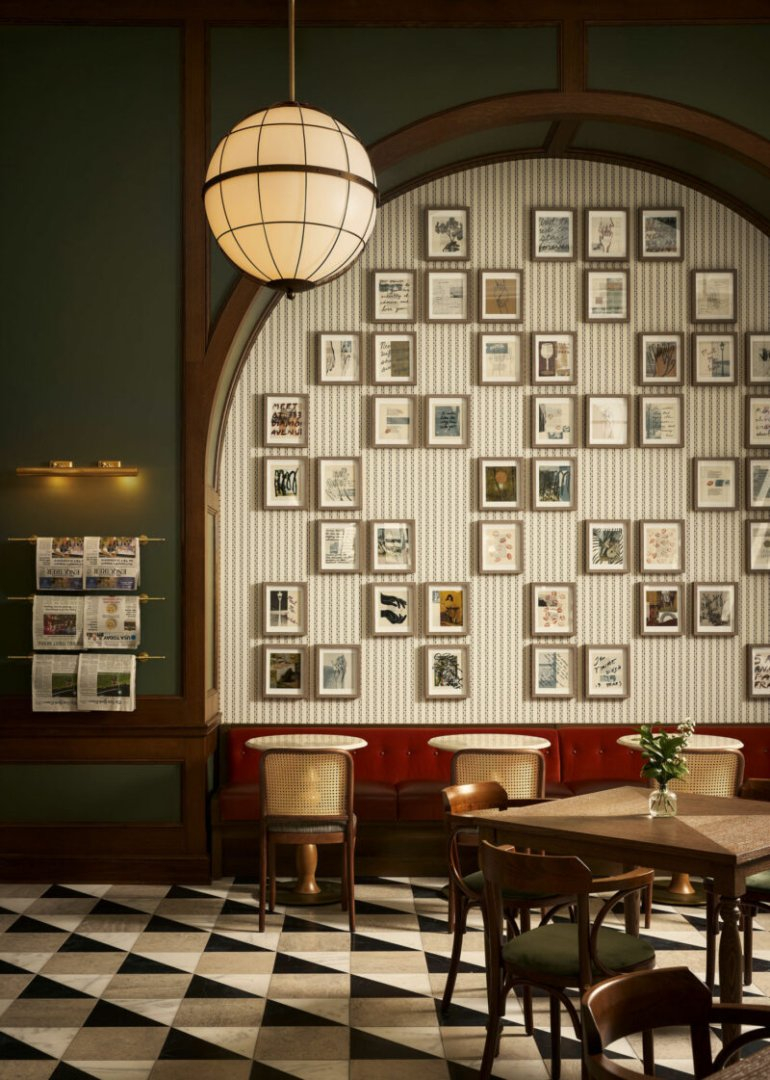 A stylish cafe with checkered floors, a red cushioned bench, wooden chairs, a large round light, and a wall covered in framed pictures and newspapers.