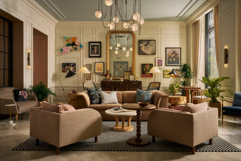 Elegant living room with four beige armchairs around a circular table, eclectic wall art, large mirror, chandelier, plants, and tall windows letting in natural light.