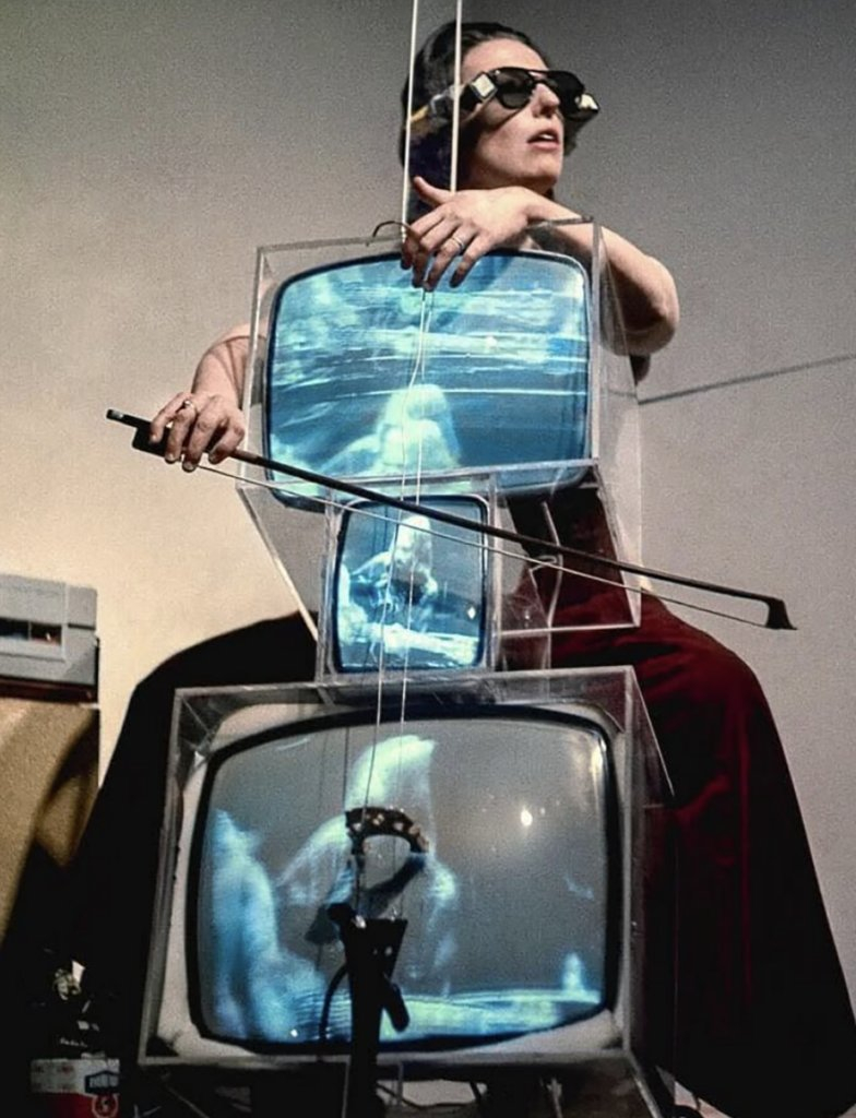 Une violoncelliste joue de son instrument par-dessus trois gros écrans de télévisions cathodiques.