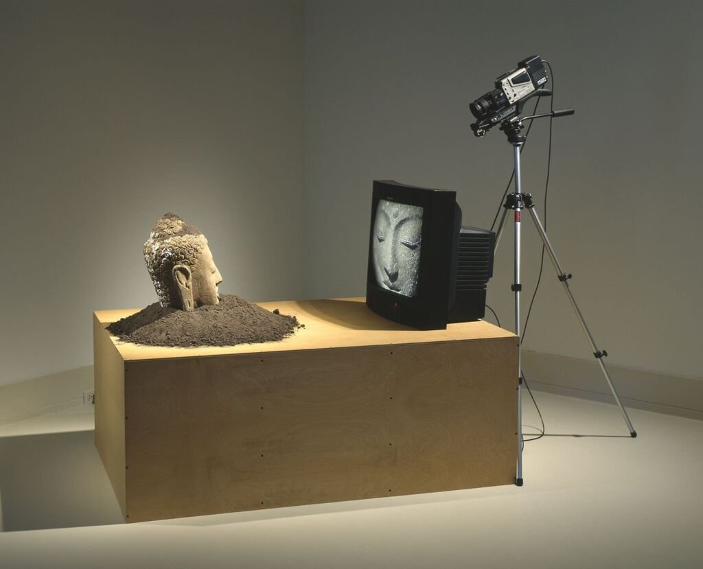 Installation artistique montrant une tête de Bouddha face à une télévision qui projette son visage.